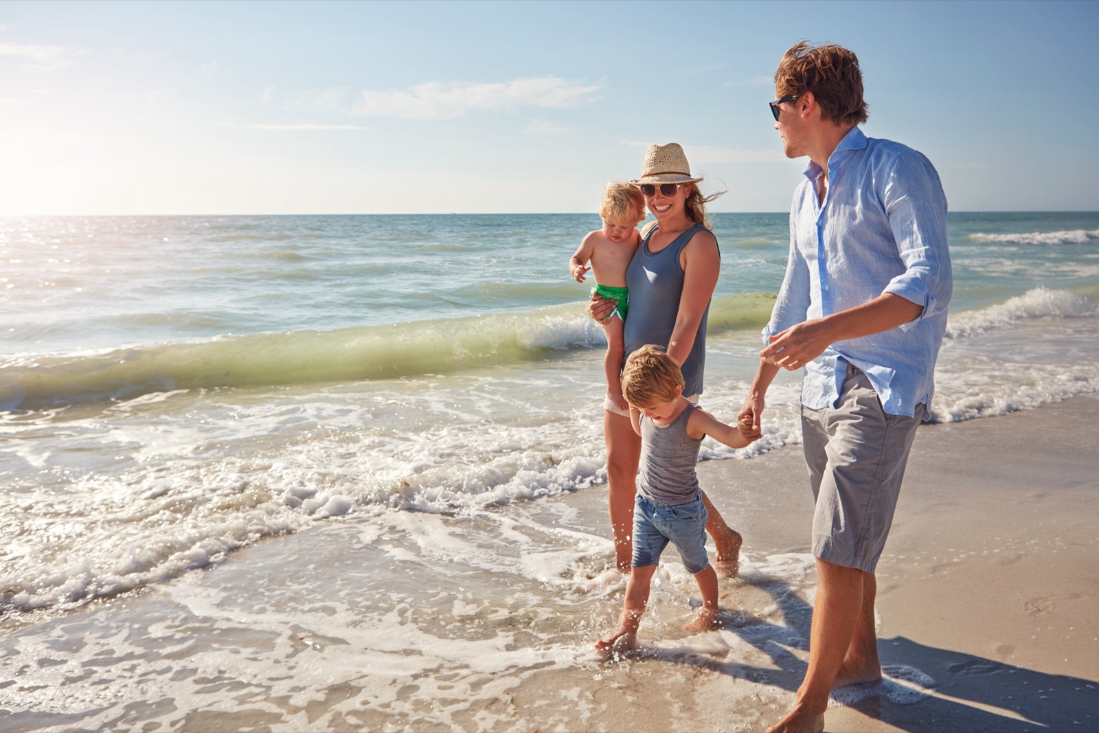 Two adults walking at the water's edge with two young children, one carried and one holding an adult's hand