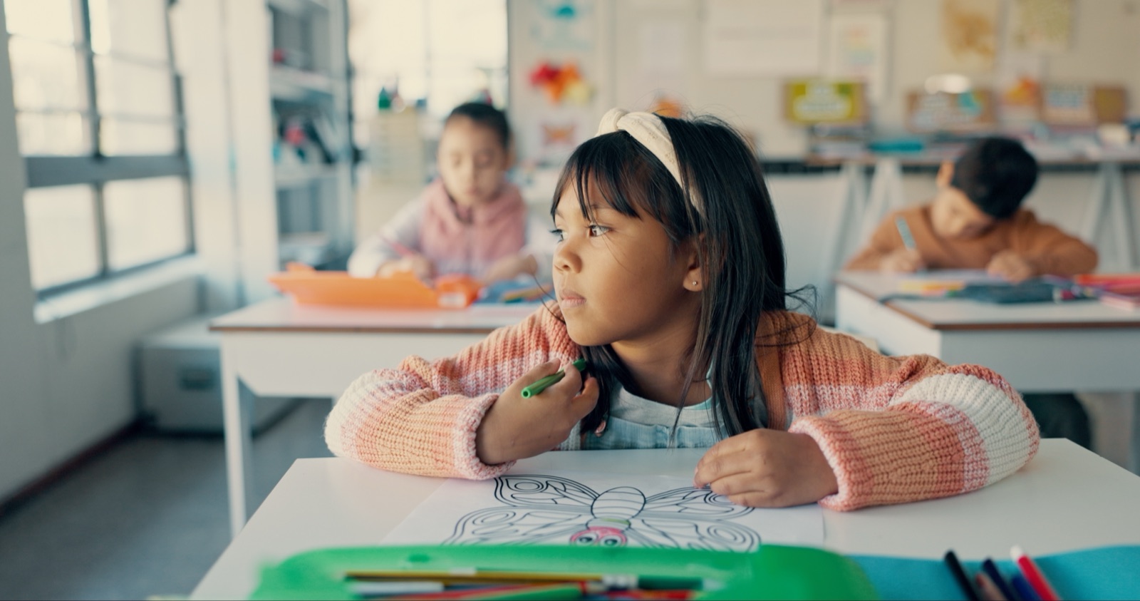 A child at a classroom desk holding a pen over a worksheet, looking away from the page