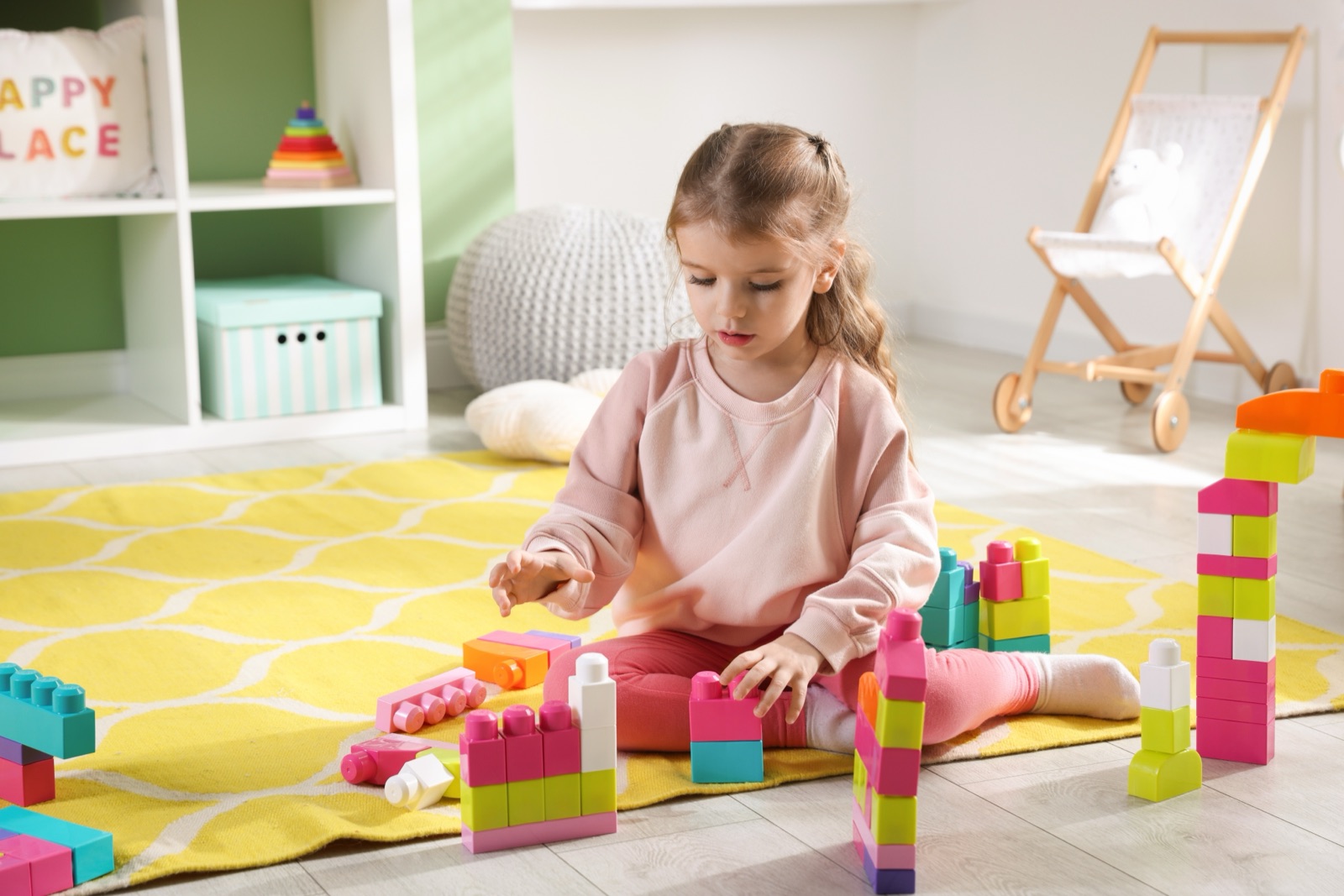 A young girl sitting alone on a rug in a bright room, absorbed in building a tower from colored blocks