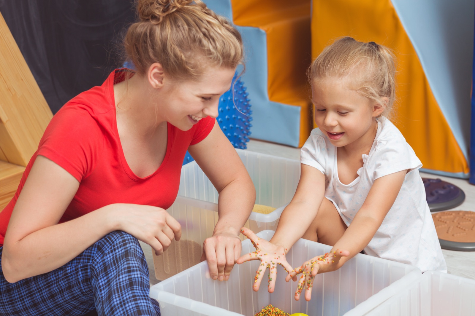 An adult and a young child with their hands in a bin of coloured dried pasta, both smiling