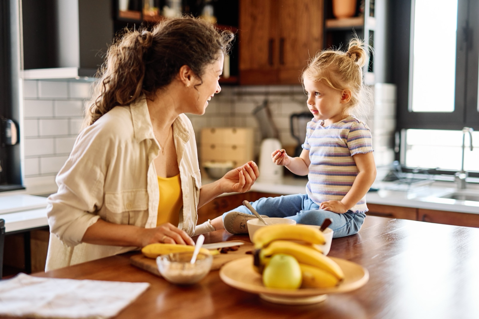 A parent and a toddler at a kitchen counter with a bowl of fruit between them