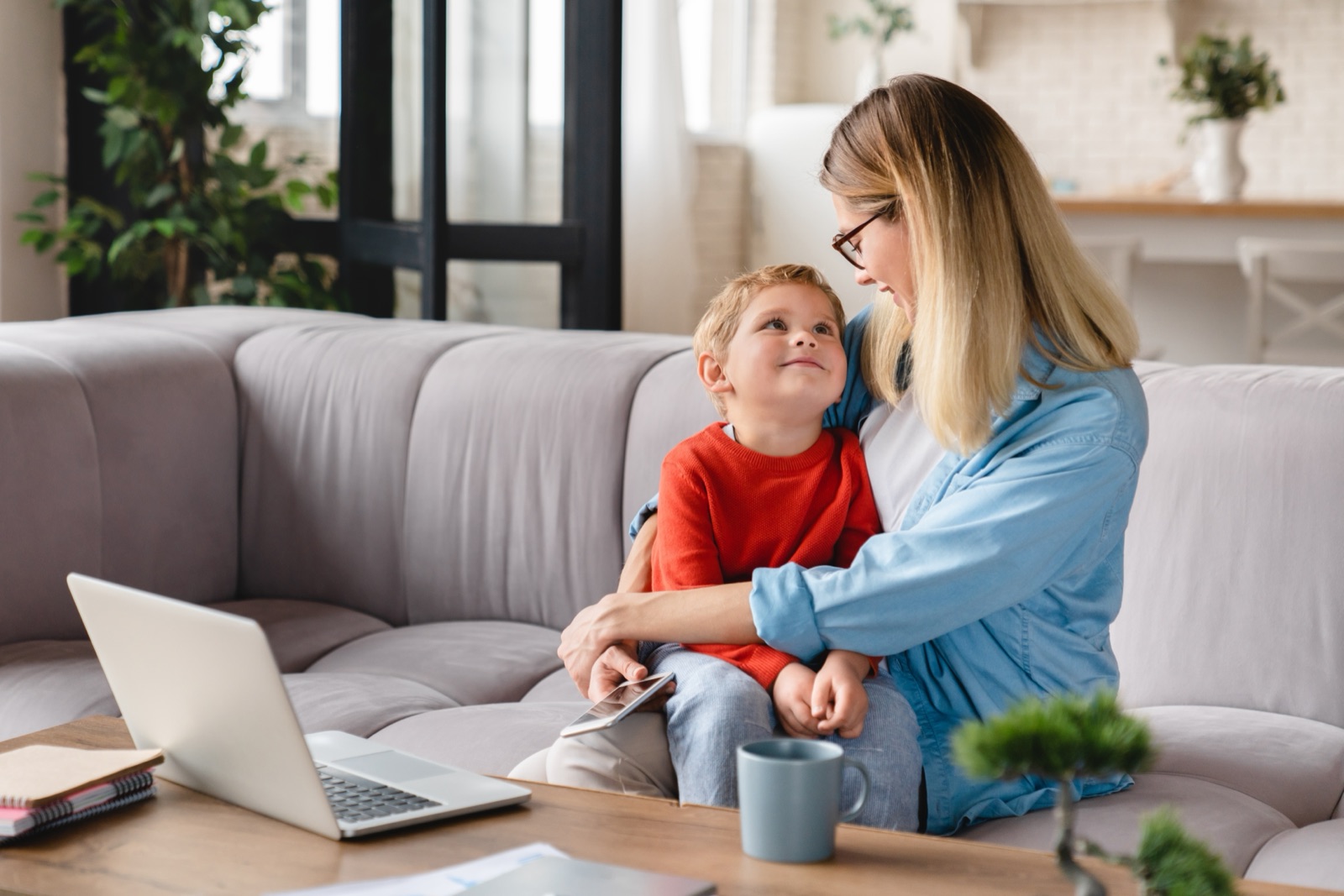A parent sitting with a young child on a sofa, a laptop and phone on the table beside them