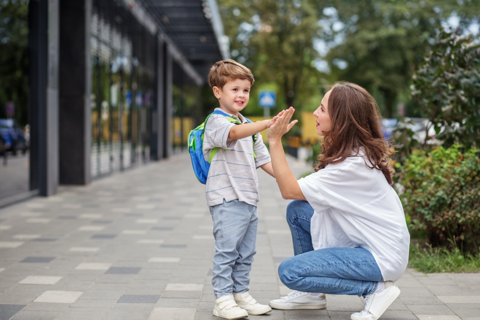 A parent crouched to a child's eye level outside a school, high-fiving them